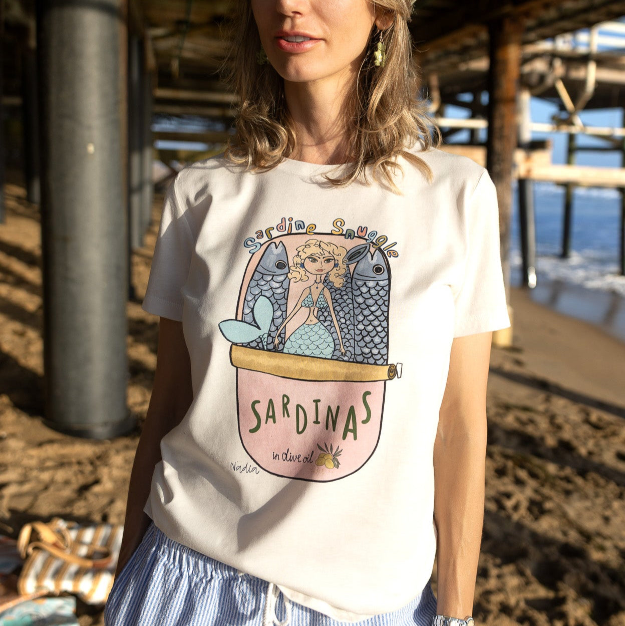 Woman standing on a beach wearing a North Shore Girls cream-colored tee with a mermaid sardine tin illustration.