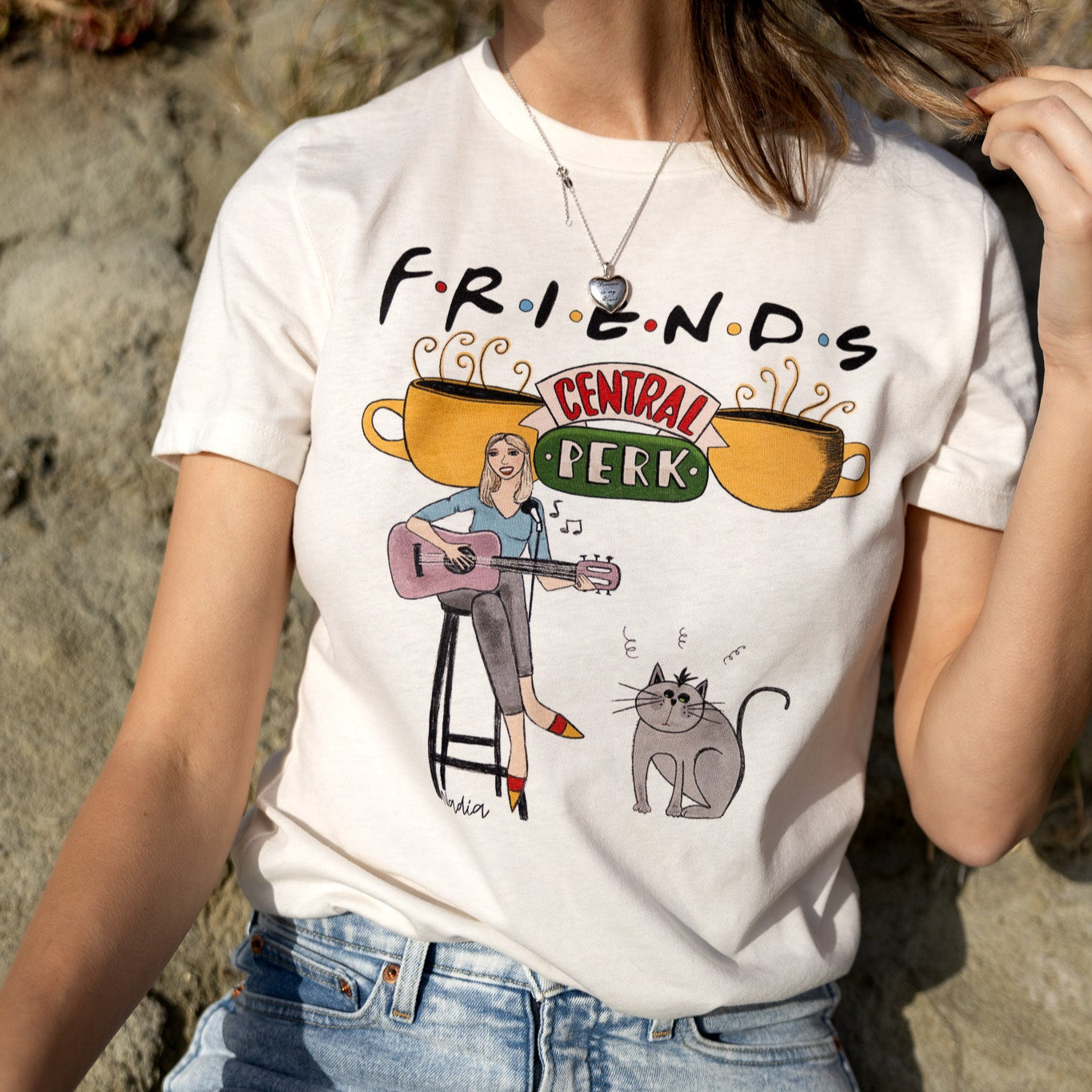 Woman wearing a white North Shore Girls tee with a hand-drawn illustration of a woman playing guitar and a "Smelly Cat"