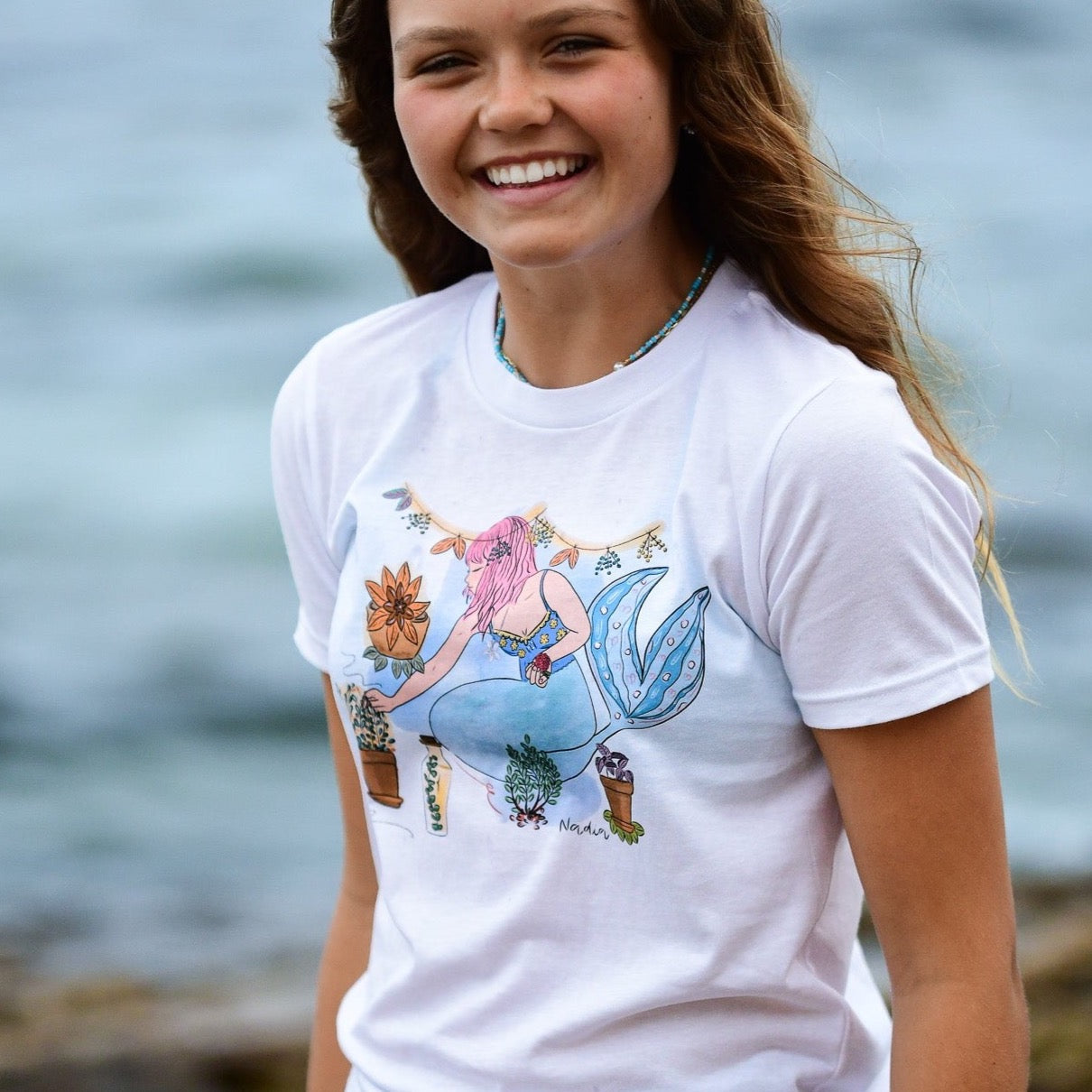 Youth white t-shirt with a hand-drawn mermaid tending to bohemian potted garden plants, North Shore Girls brand.