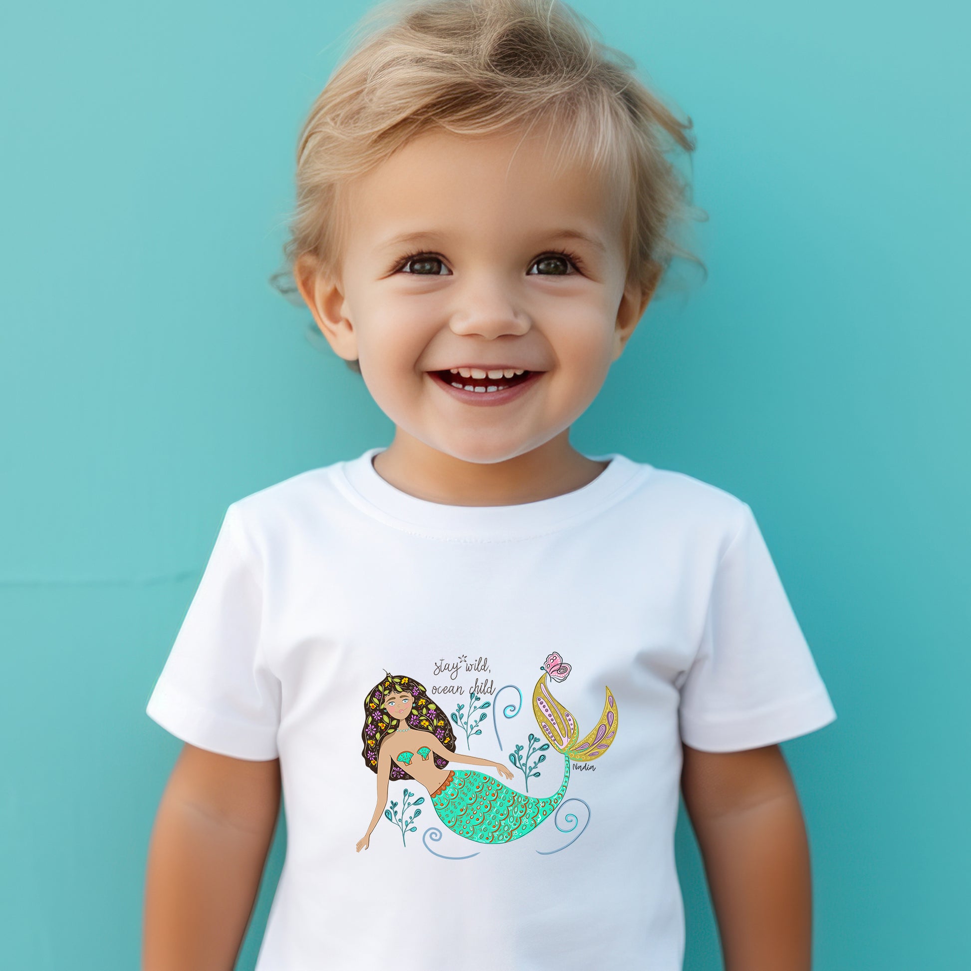 Toddler wearing a white tee with a colorful mermaid illustration and “Stay Wild Ocean Child” slogan – original artwork by Nadia Watts