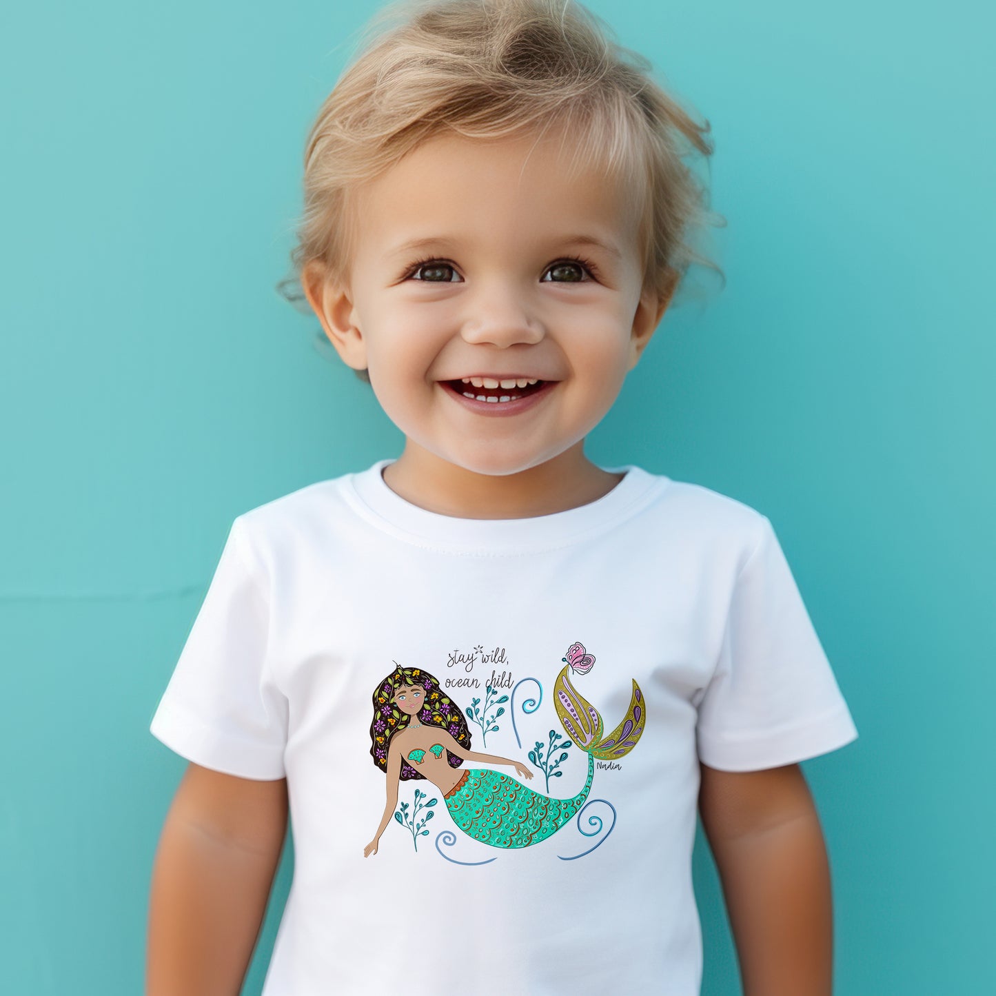 White toddler t-shirt featuring a hand-drawn mermaid with flowers in her hair and "Stay Wild Ocean Child" text, signed by the artist