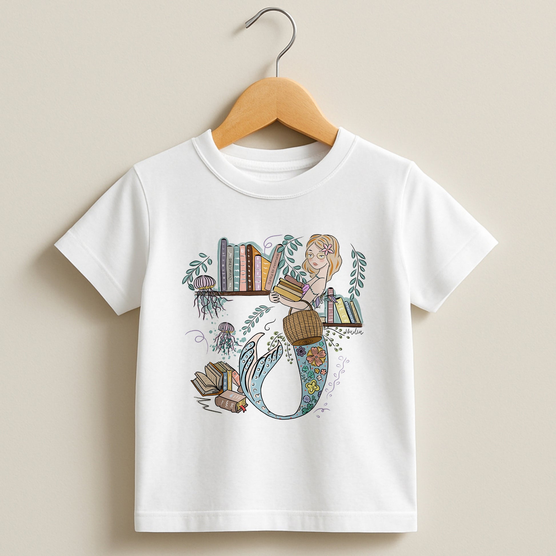 White toddler t-shirt on a wooden hanger featuring a hand-illustrated mermaid librarian with books and jellyfish, signed by the artist.