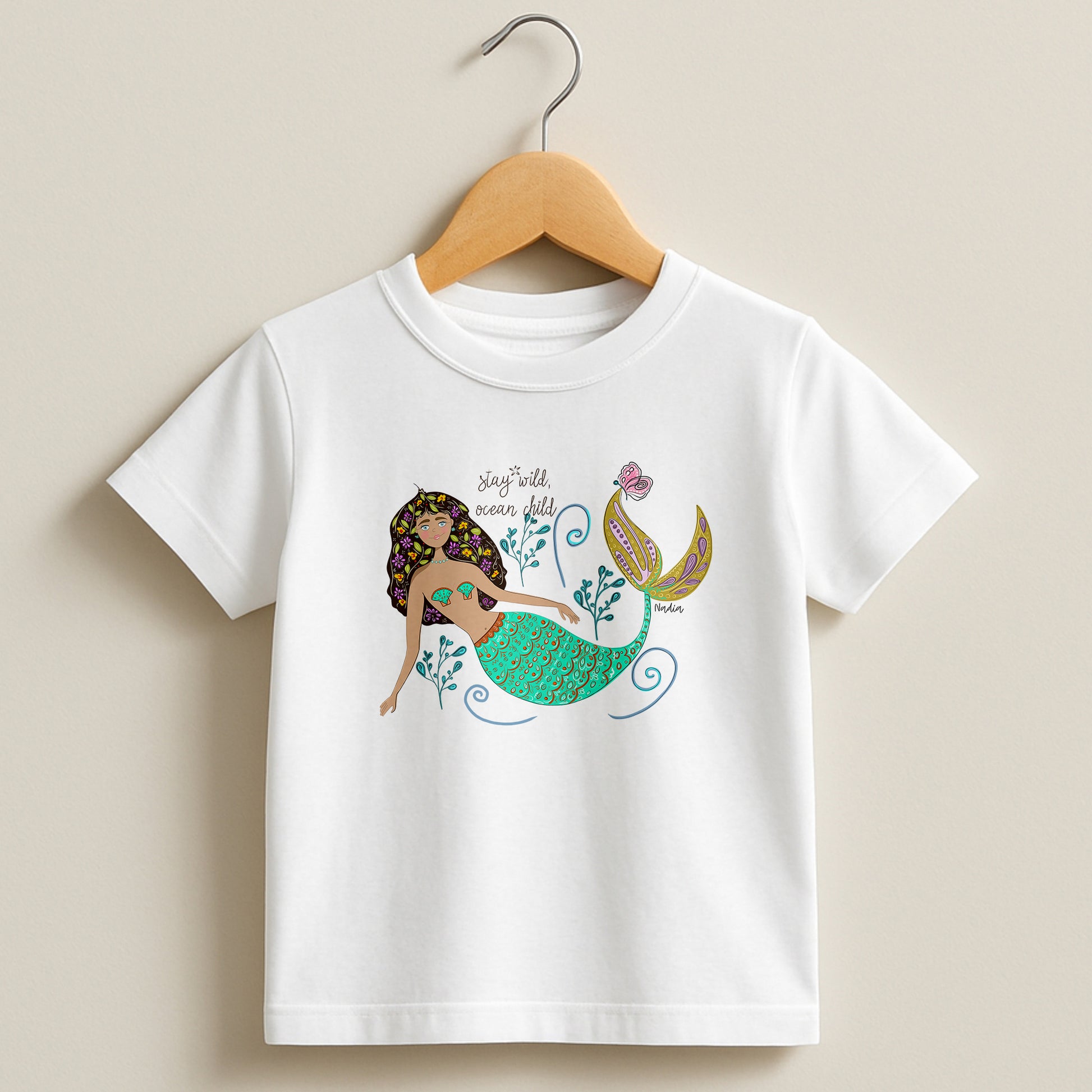 White toddler t-shirt featuring a hand-drawn mermaid with flowers in her hair and "Stay Wild Ocean Child" text, signed by the artist